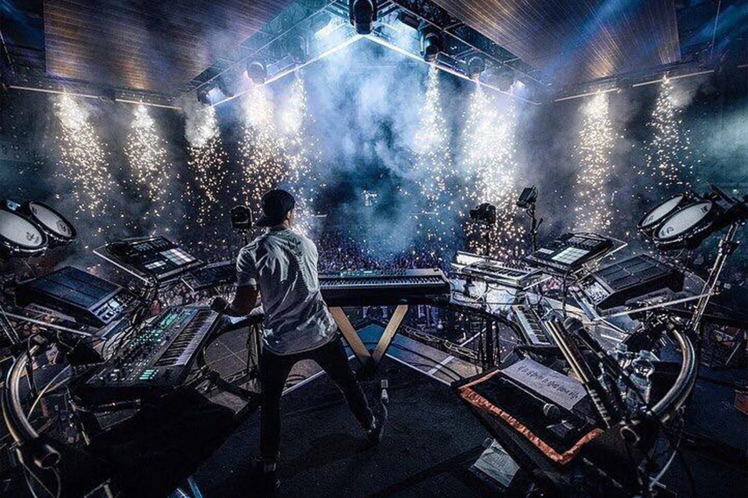 A DJ performing on stage at a concert with sparklers and sparks shooting into the air, a large crowd in the background, and DJ equipment including turntables, mixers, and keyboards surrounding the DJ.