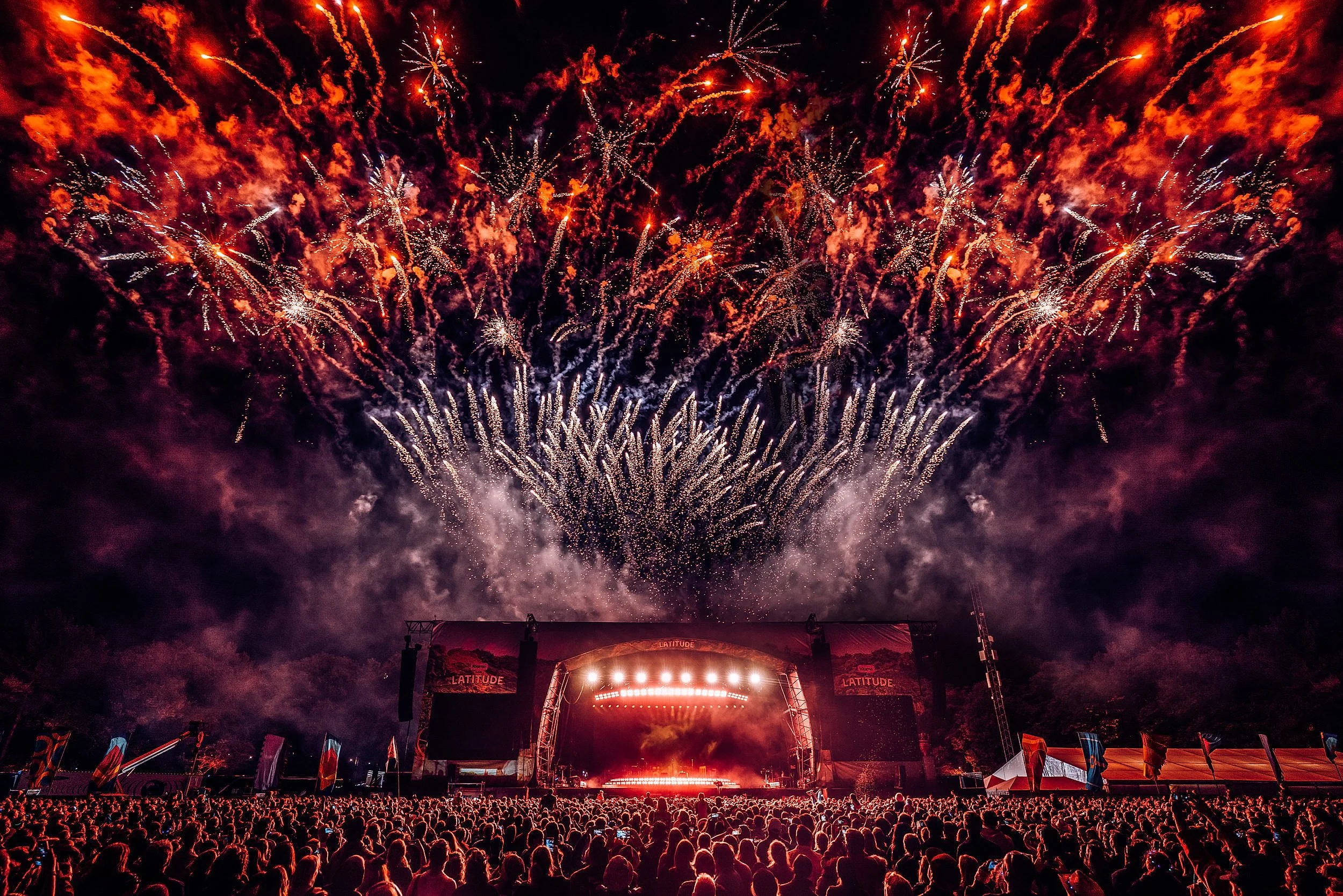 Fireworks illuminate the night sky over a large crowd attending an outdoor concert, with a stage and bright lights at the bottom of the scene.