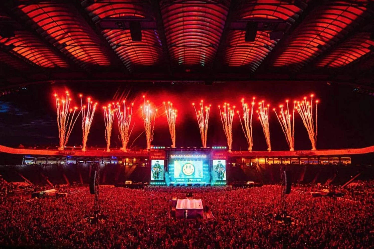 Fireworks display at a large outdoor concert or stadium with a stage, screens, and a cheering crowd under a curved roof structure.