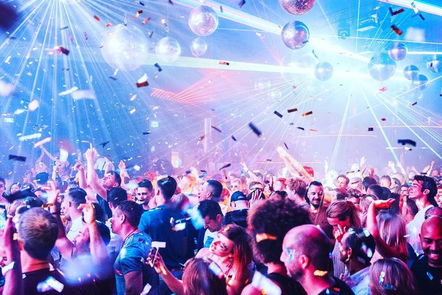 A lively party scene with a large crowd of people dancing and celebrating under colorful lights and disco balls, with confetti falling from above.