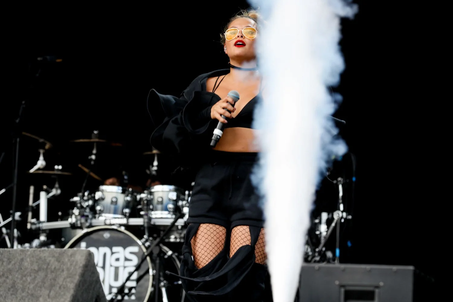 A female singer with blonde hair, wearing yellow sunglasses, a black crop top, black shorts, fishnet stockings, and a black jacket draped over her shoulders, performing on stage holding a microphone. A drum set is visible in the background, and there is a white smoke or vapor in the foreground.