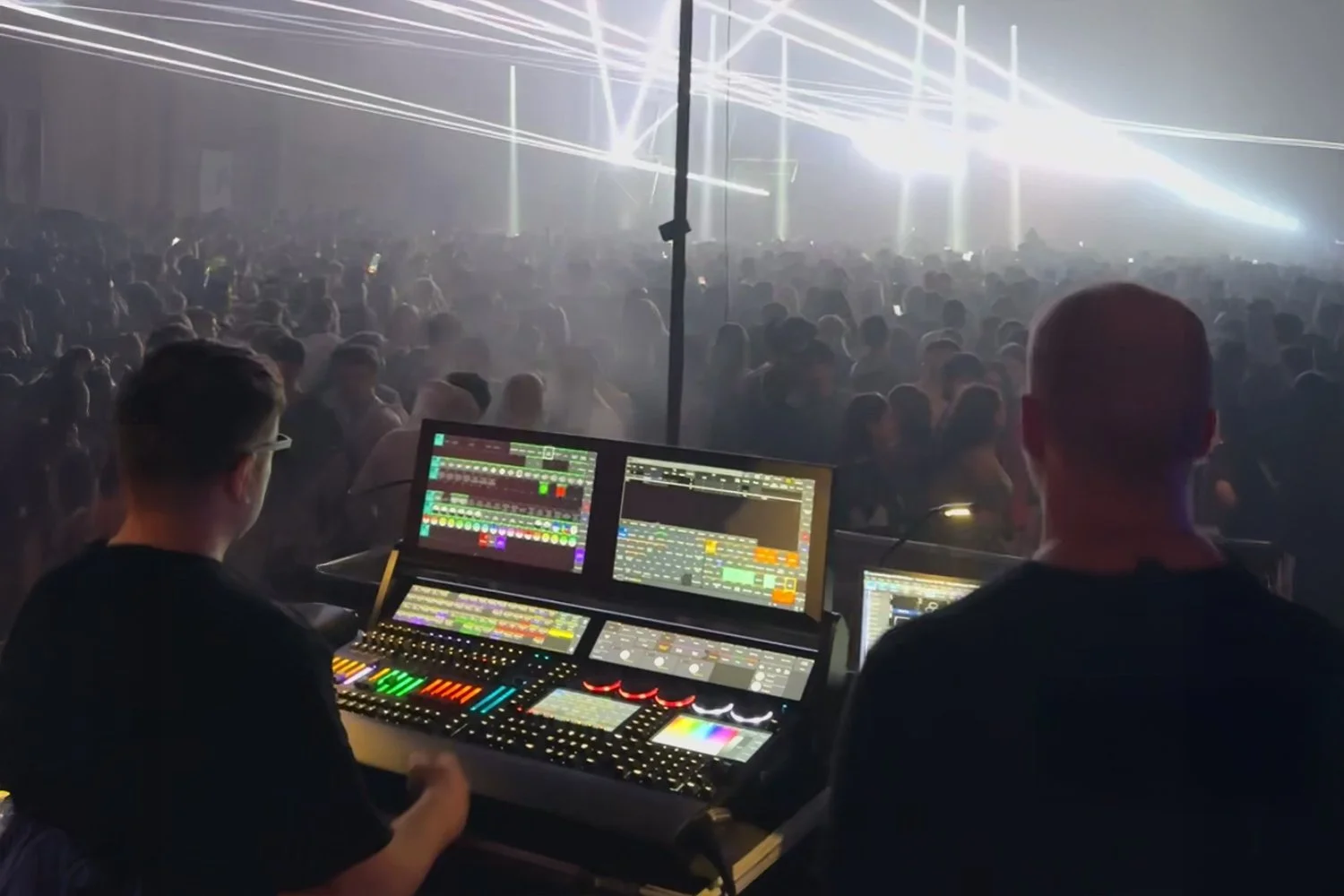 Two sound engineers operating a mixing console at a music concert, with a large crowd and laser lights in the background.