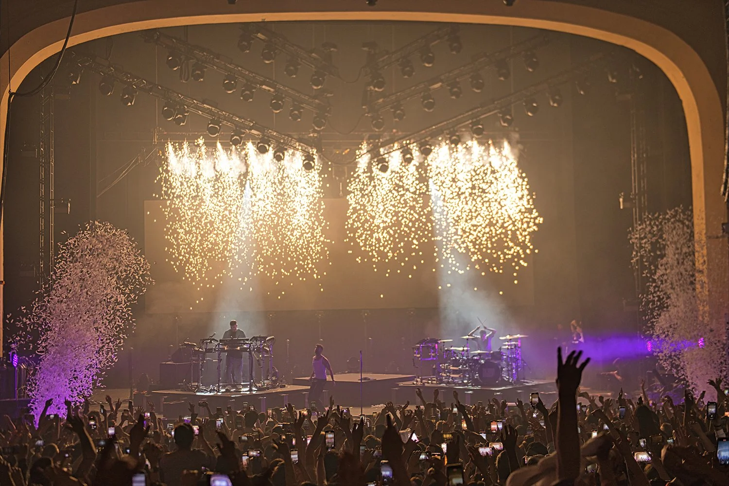 A live concert with a band performing on stage, fireworks, and confetti in the air. The audience is visible with many people holding up their phones.
