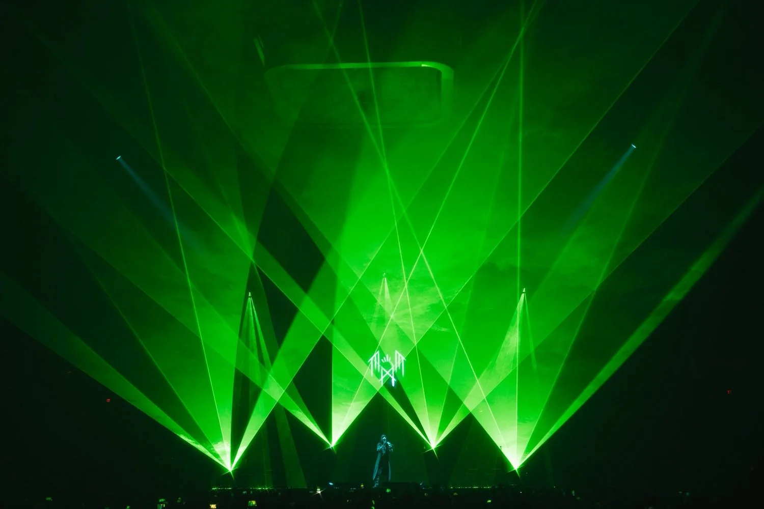 Green laser lights projecting in a concert or event scene with a silhouette of a person standing on stage in front of a large, abstract structure.