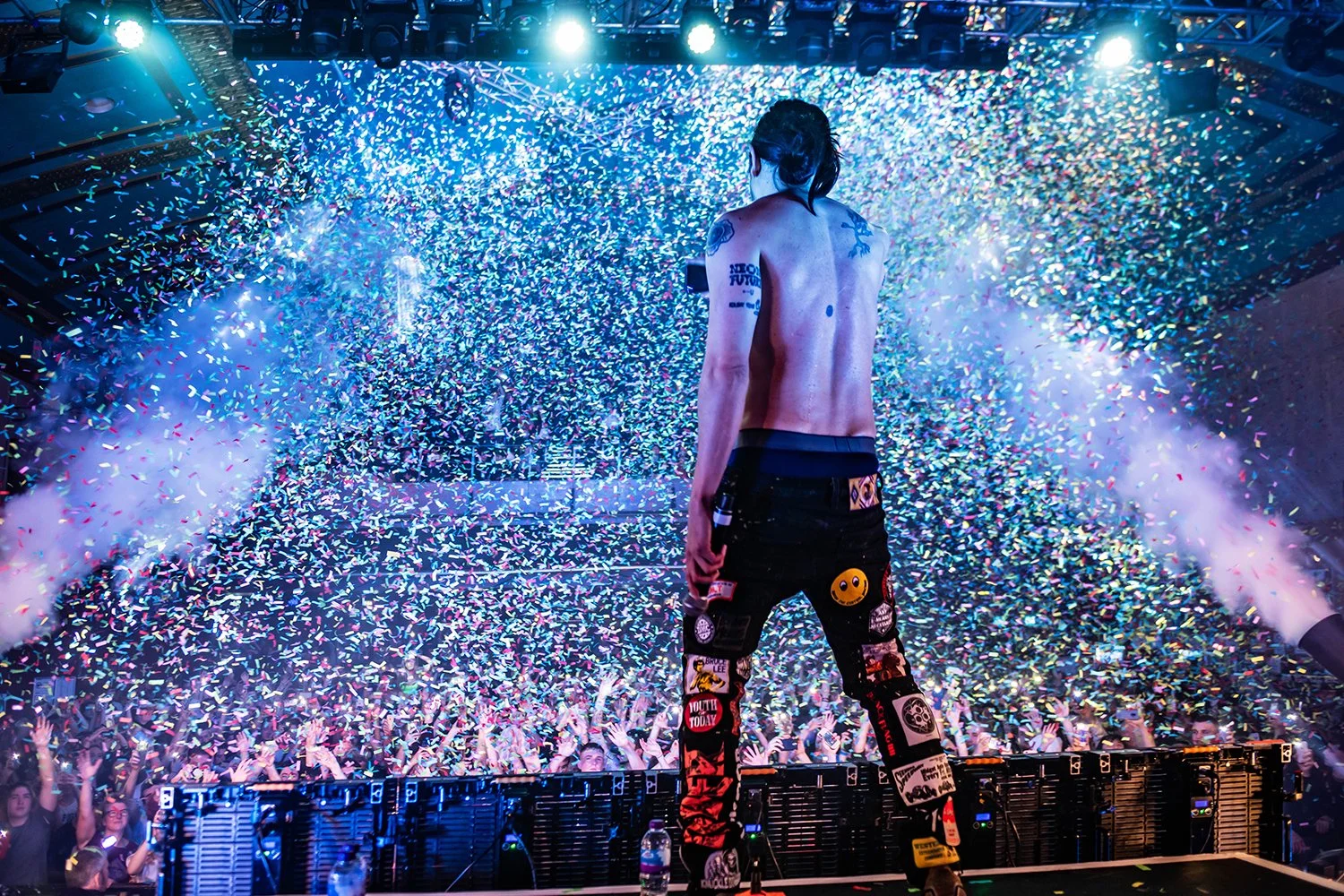 A performer on stage facing a large crowd, with colorful confetti falling all around, illuminating the scene with bright lights.