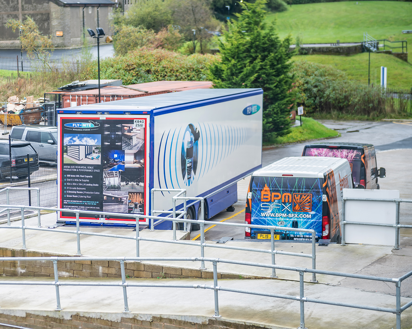 View of parked trucks in a lot, including a large white trailer with advertising for a rehearsal studio and a smaller van with graffiti-style branding, behind a metal railing.