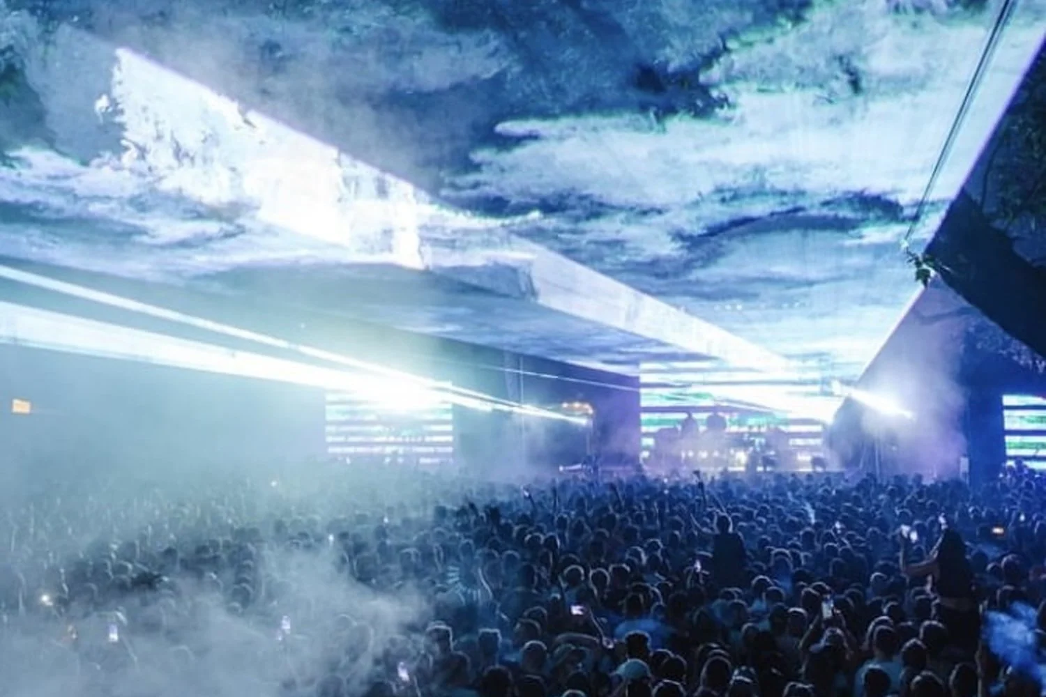 Large indoor concert with a crowd of people, bright stage lights, and a sky-like ceiling projection.