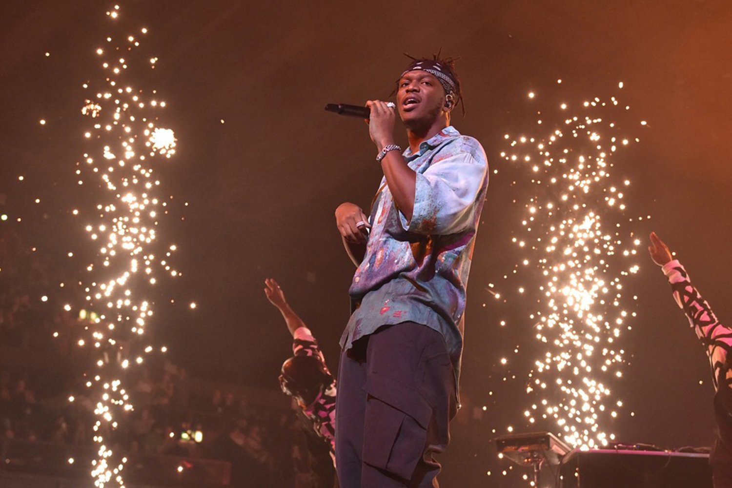 A male performer holding a microphone on stage with sparks flying in the background during a concert.