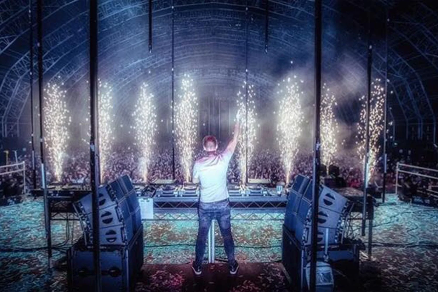 DJ standing with one arm raised in front of a large crowd at a concert, with bright fireworks and smoke effects behind them.
