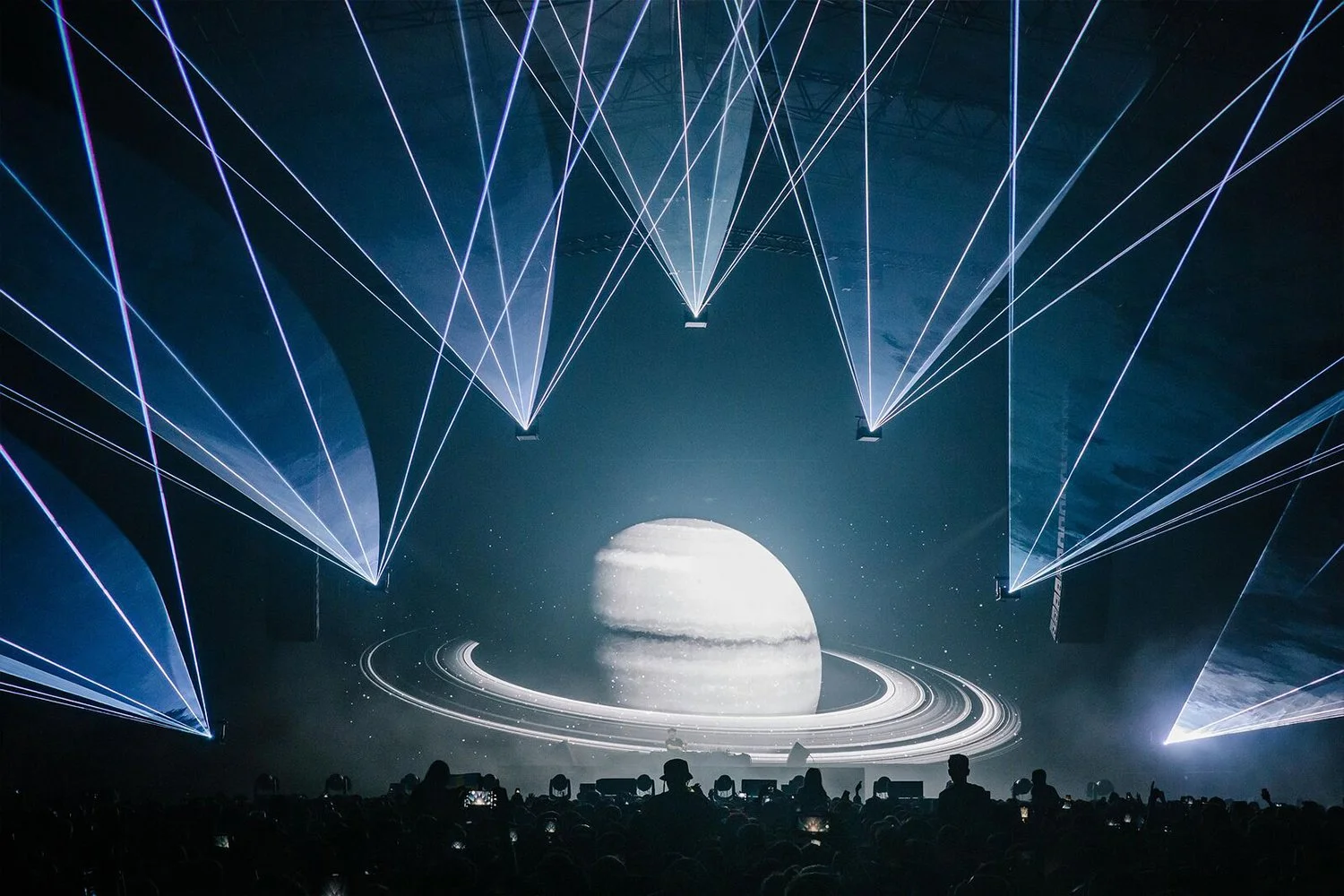 A concert or event with stage lighting and a large projection of a planet with rings, resembling Saturn, in the background.