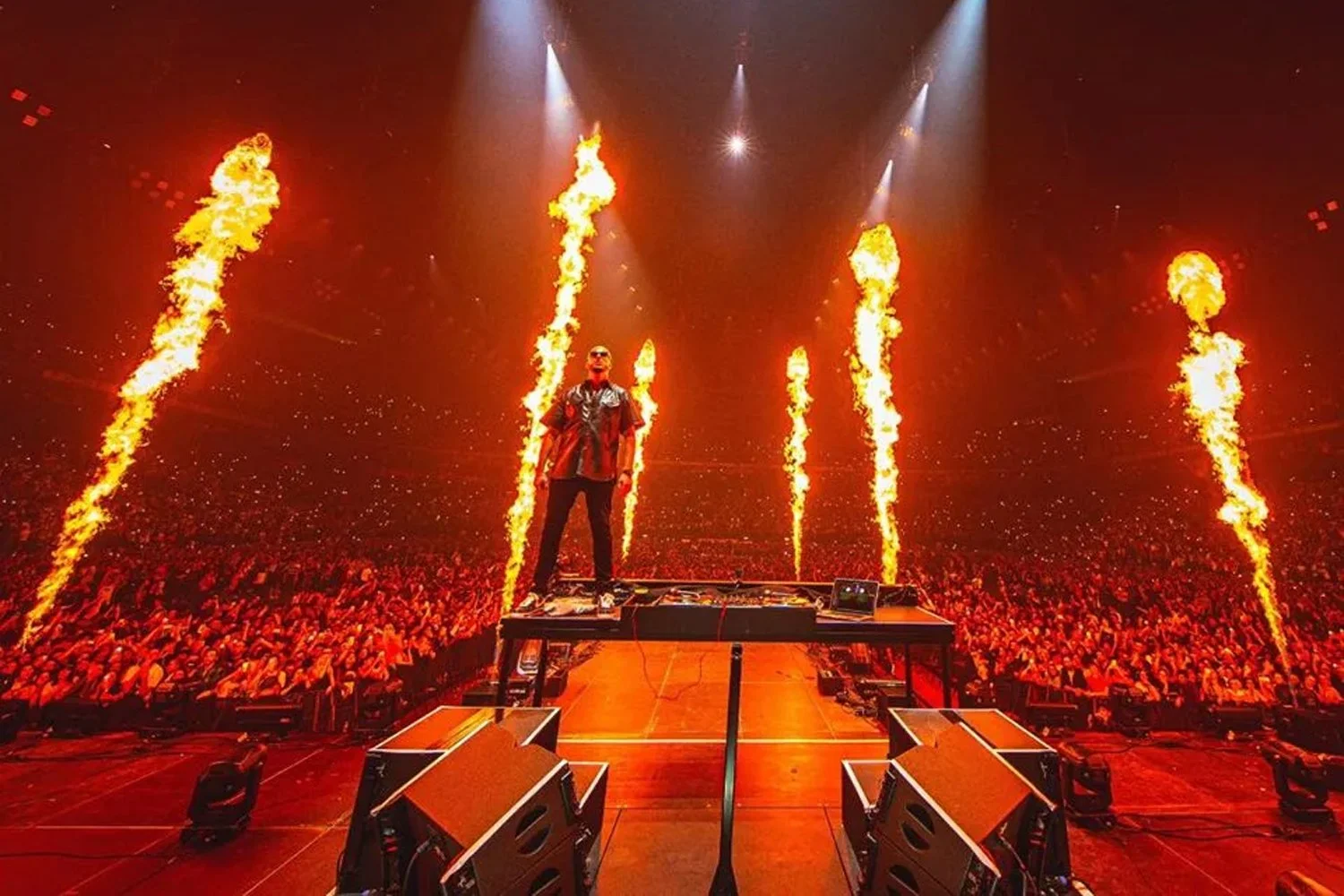 A performer stands on a stage with fire effects shooting from behind him during a concert, with a large crowd in the background.