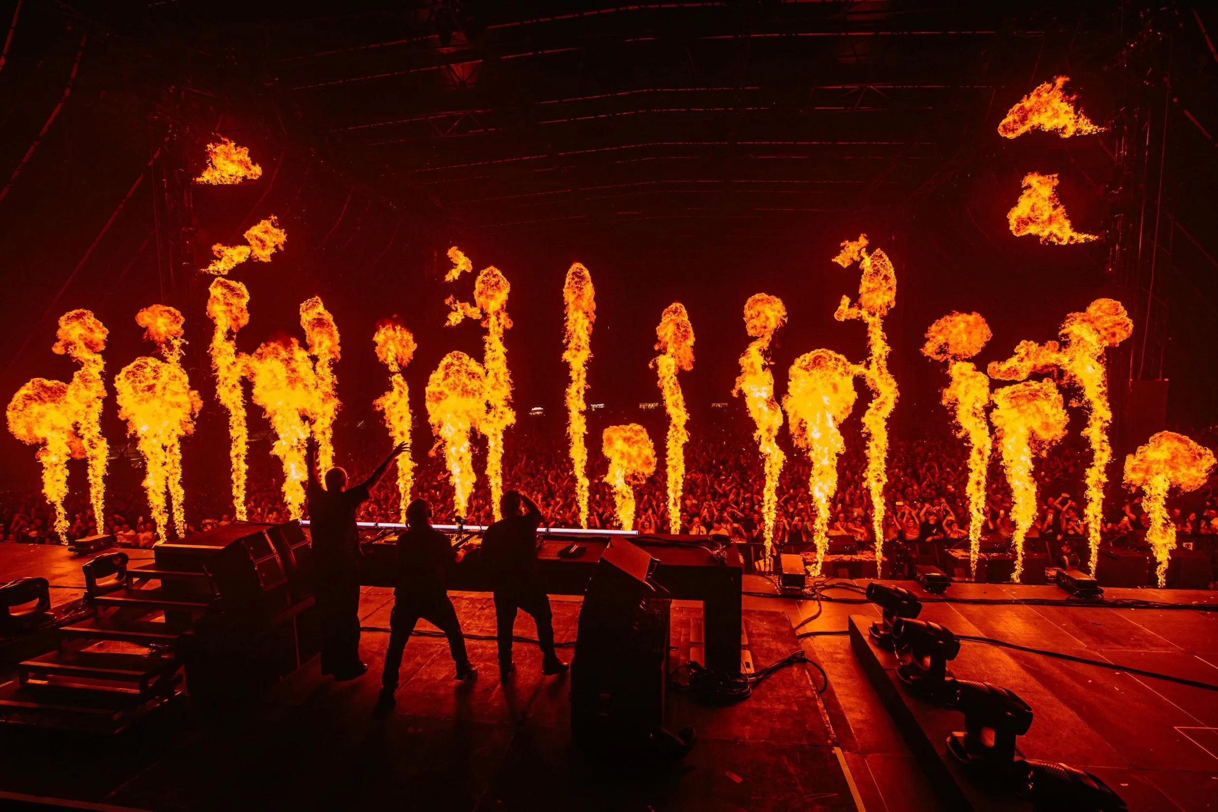 A concert stage with fire effects shooting flames into the air, with silhouettes of performers and a large crowd in the background.