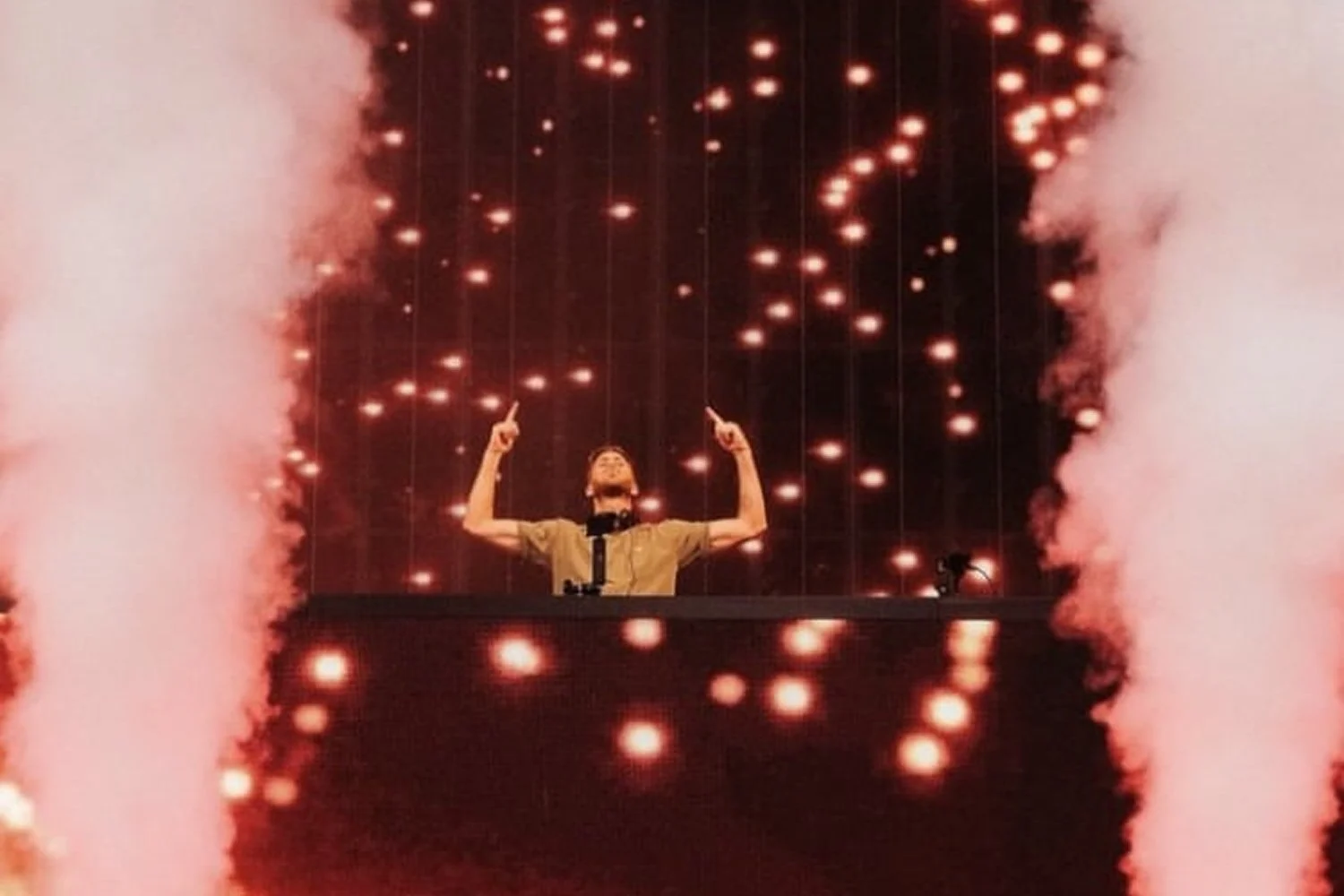 A person standing behind a DJ booth with arms raised, surrounded by pink smoke and falling orange sparks, with a dark background.