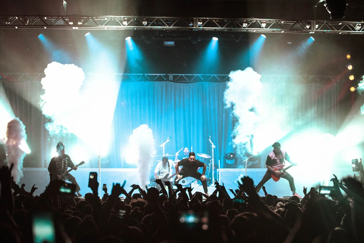 A live concert scene with a band performing on stage, surrounded by bright lights, smoke effects, and an audience with raised hands.