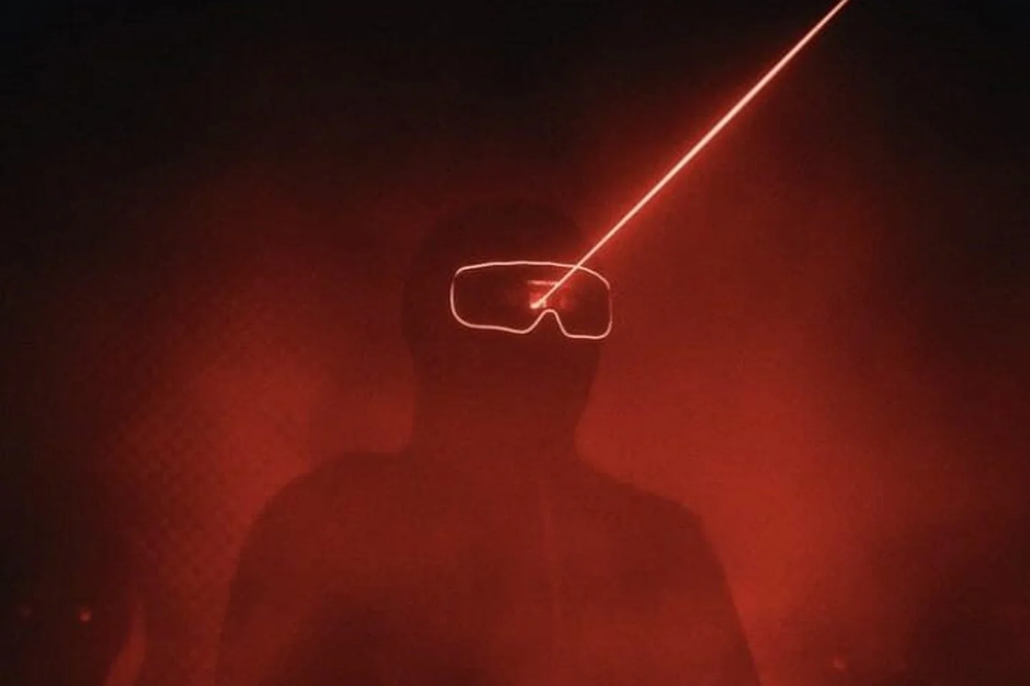 Person wearing goggles in a dark room, with a red laser beam directed at the goggles, creating a glowing outline around the glasses and a beam extending outward.