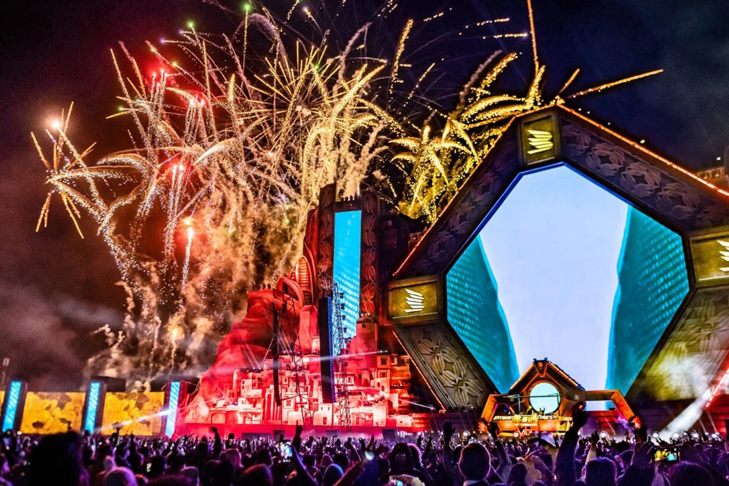 Fireworks display over a large music festival stage at night with an audience watching.