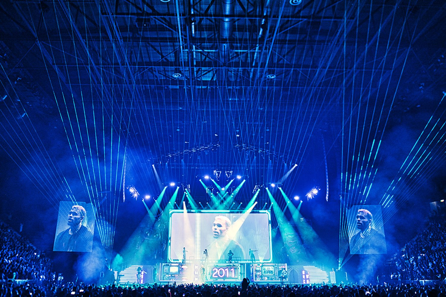 A concert stage with blue lighting, laser lights, large screens displaying a performer, and a crowd of attendees.