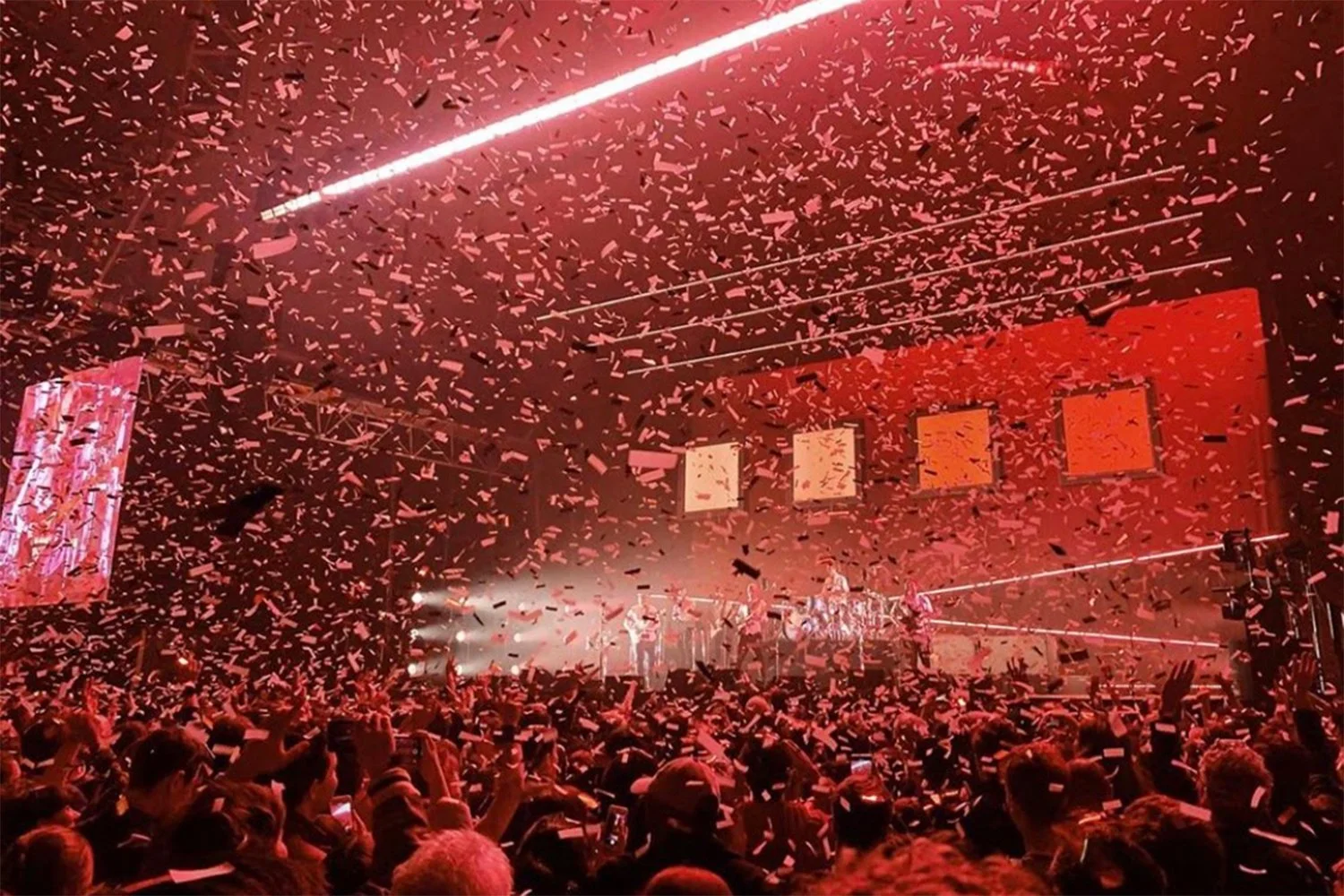 A concert scene with a large crowd, red lighting, laser graphics, and confetti falling from above.