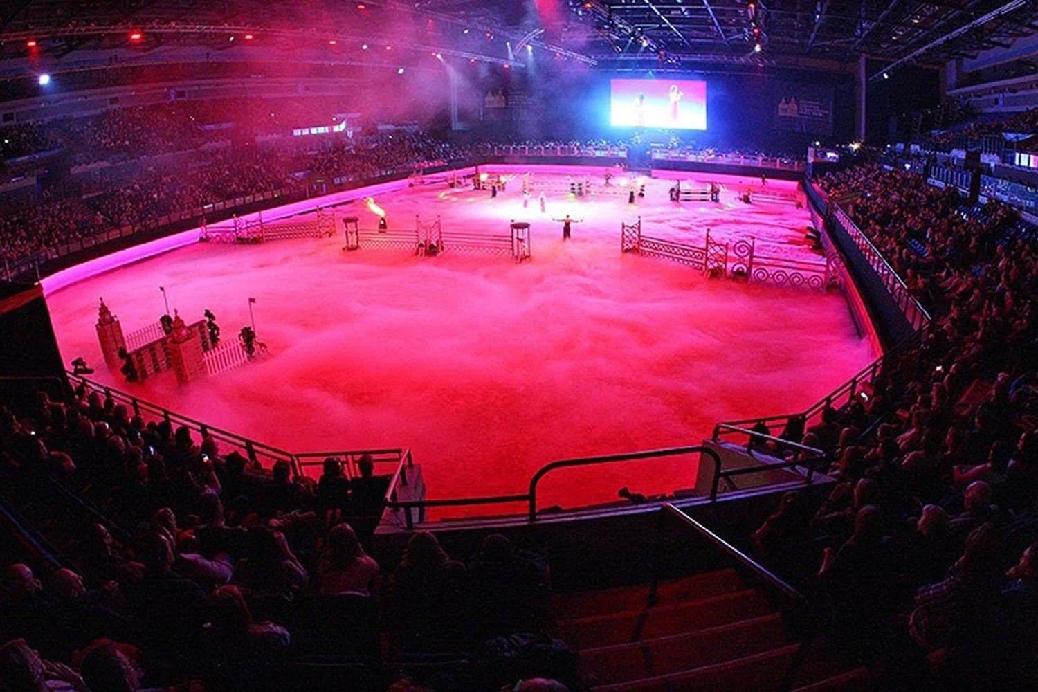 Indoor arena with pink lighting and fog, filled with spectators, set up for a dog show or competition with jumps and obstacles on the sandy floor.