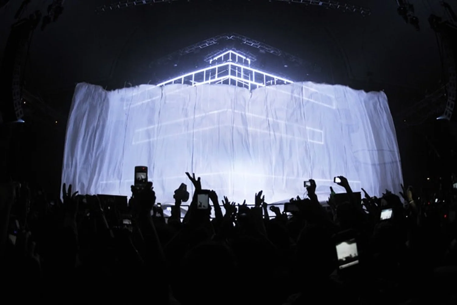 Concert scene with a large, illuminated stage covered by a white fabric and surrounded by people taking photos with smartphones.