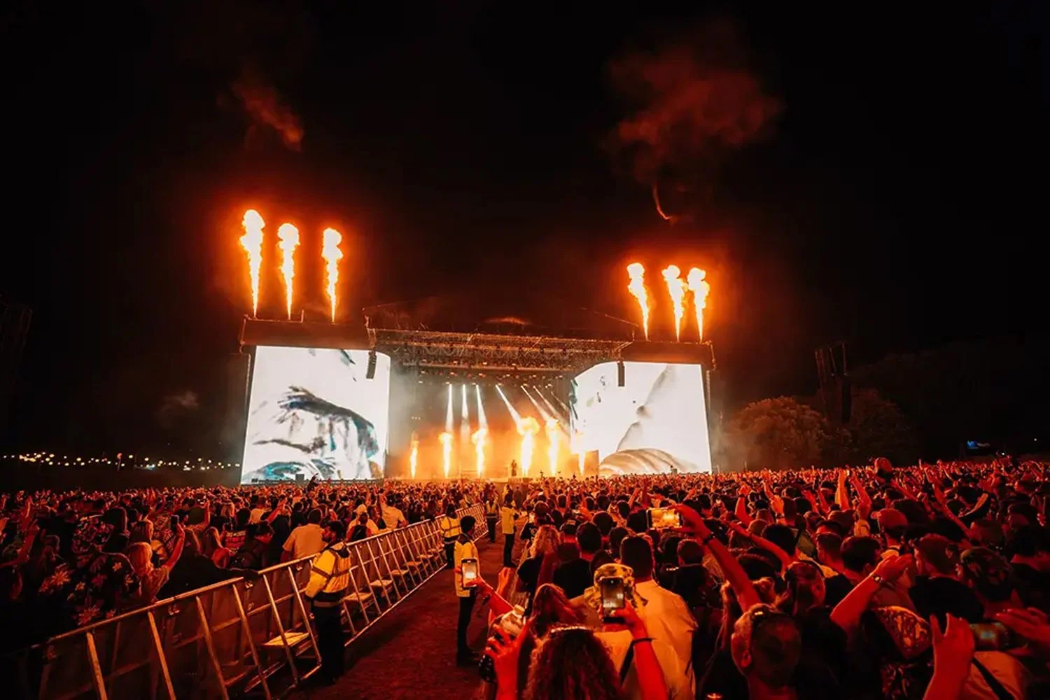 A large crowd at a nighttime outdoor concert with a stage, fireworks, and bright screens.