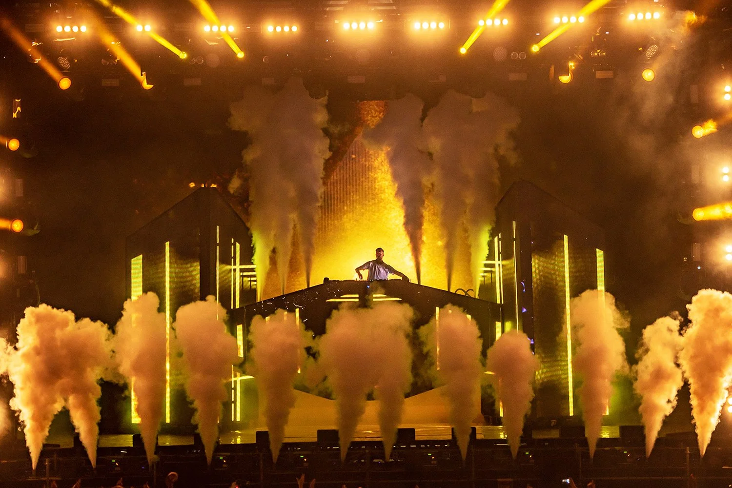 A DJ performs on stage surrounded by bright yellow lights, smoke machines, and pyrotechnics, creating an energetic concert atmosphere.