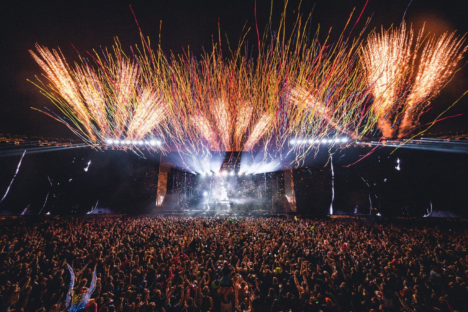 Colorful fireworks explode over a large outdoor concert with a packed crowd at night.