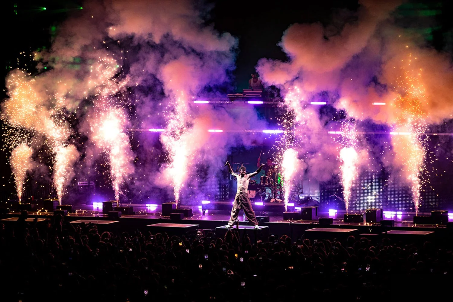 A live concert performance with a singer on stage, surrounded by colorful pyrotechnics, smoke, and bright lights, with an audience in the foreground.