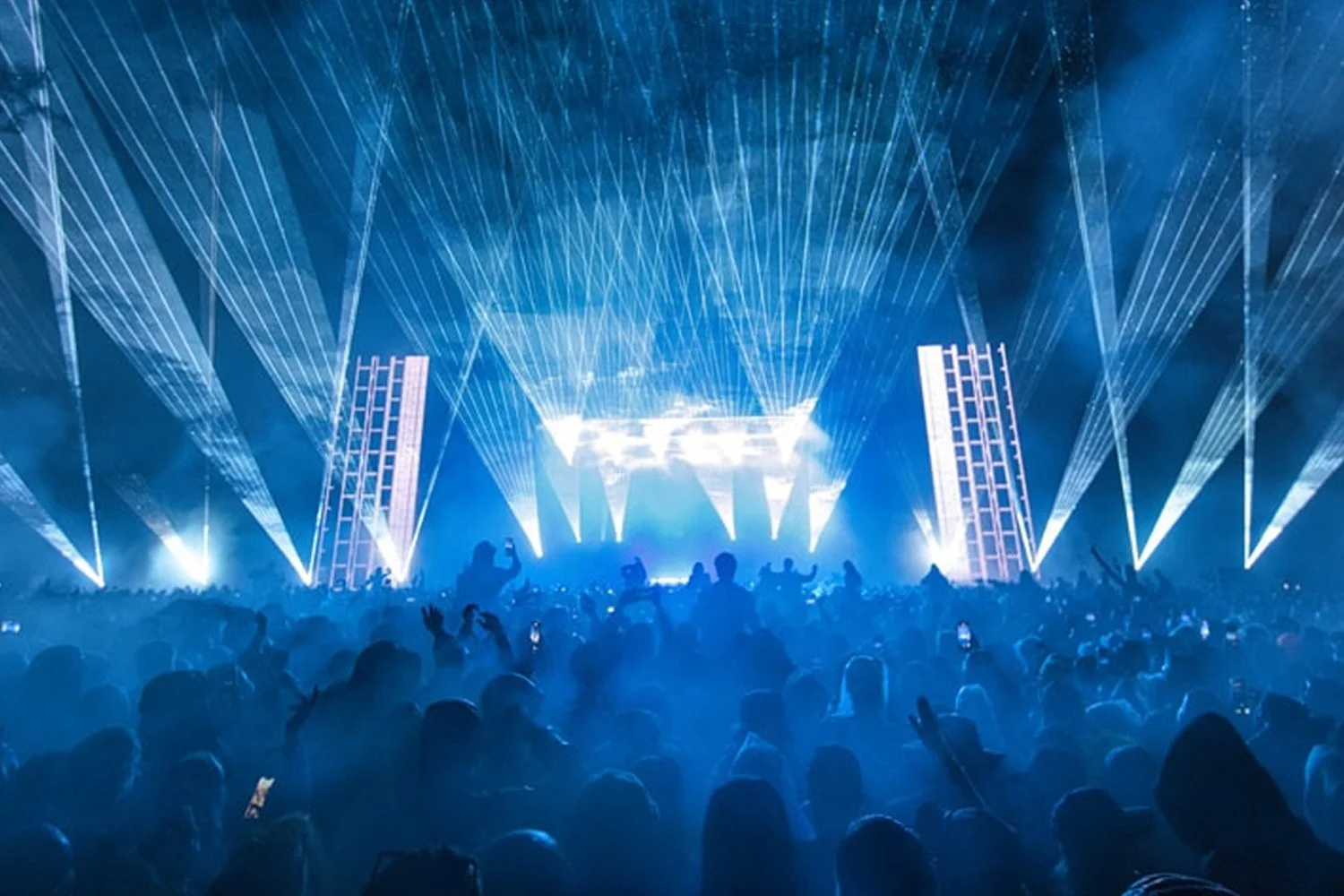 Crowd of concert attendees at a music festival with bright blue laser lights and stage in the background.