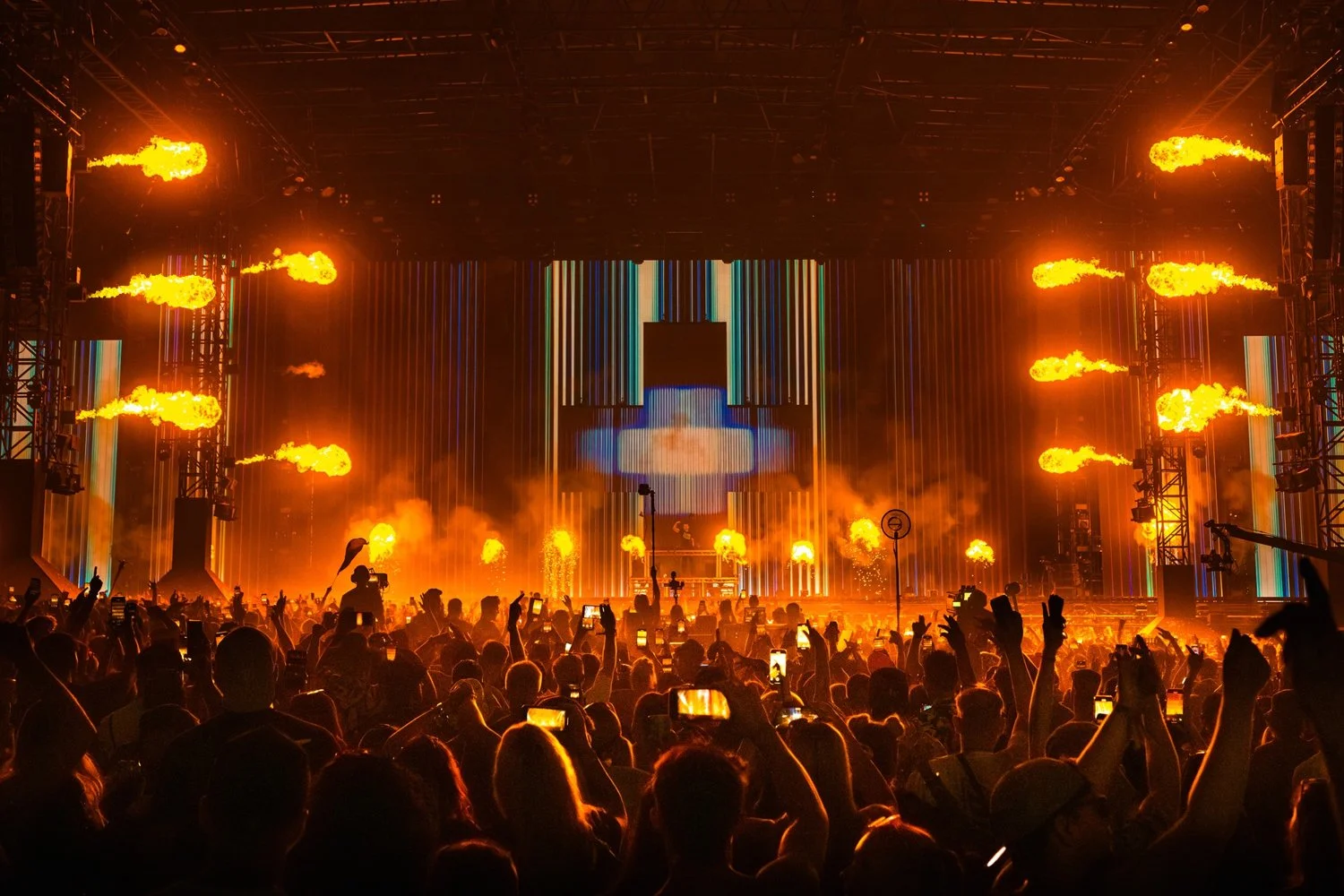 Crowd at a concert with a stage featuring fire effects, bright vertical light strips, and a large screen, during a lively music event.