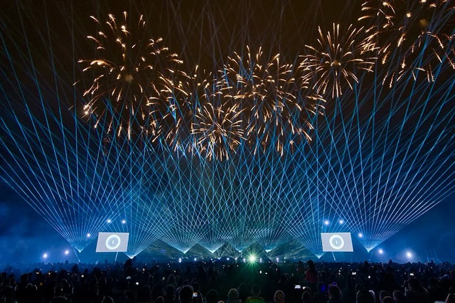 Fireworks display over a crowd at night with blue laser light beams and two large screens showing a clock icon.