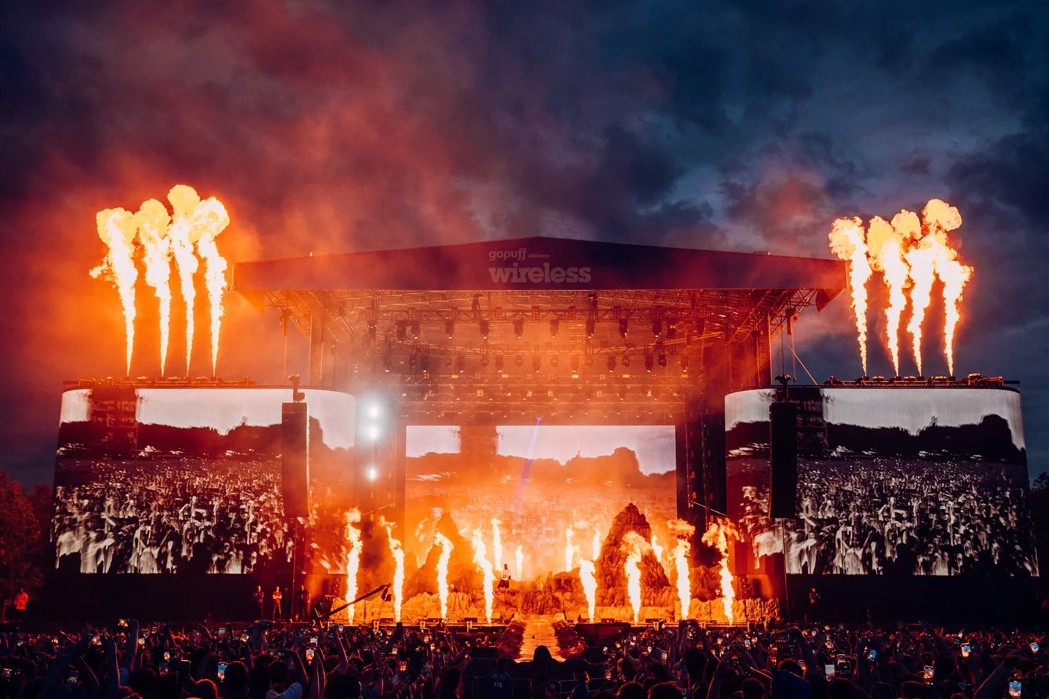A large outdoor concert stage at dusk with bright flames shooting upwards from various points on the stage, multiple large screens displaying fiery visuals, and a crowd of people taking photos and videos.