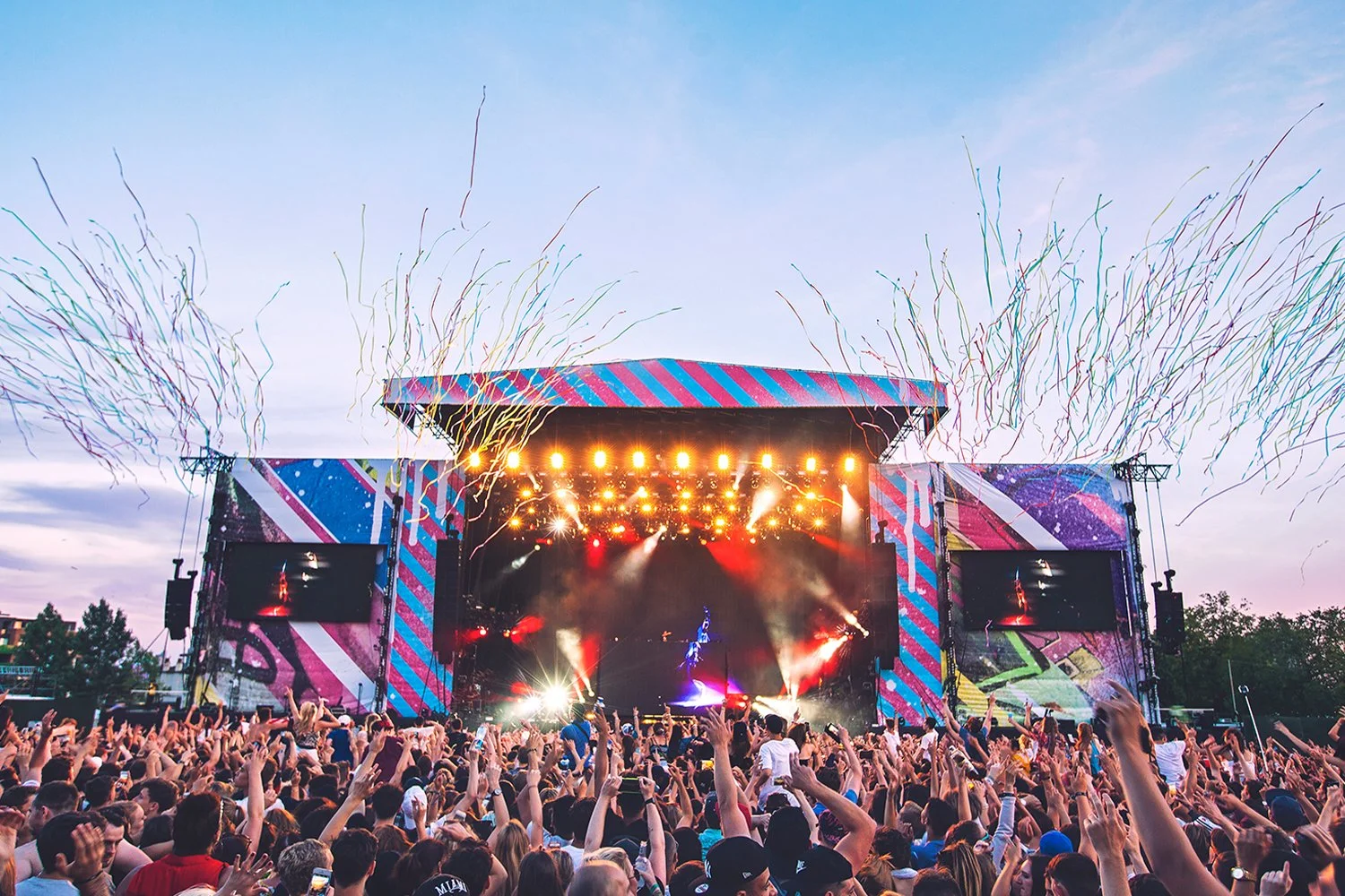 A large outdoor music concert with a crowd, a brightly lit stage, colorful decorations, and streamers in the sky during sunset.