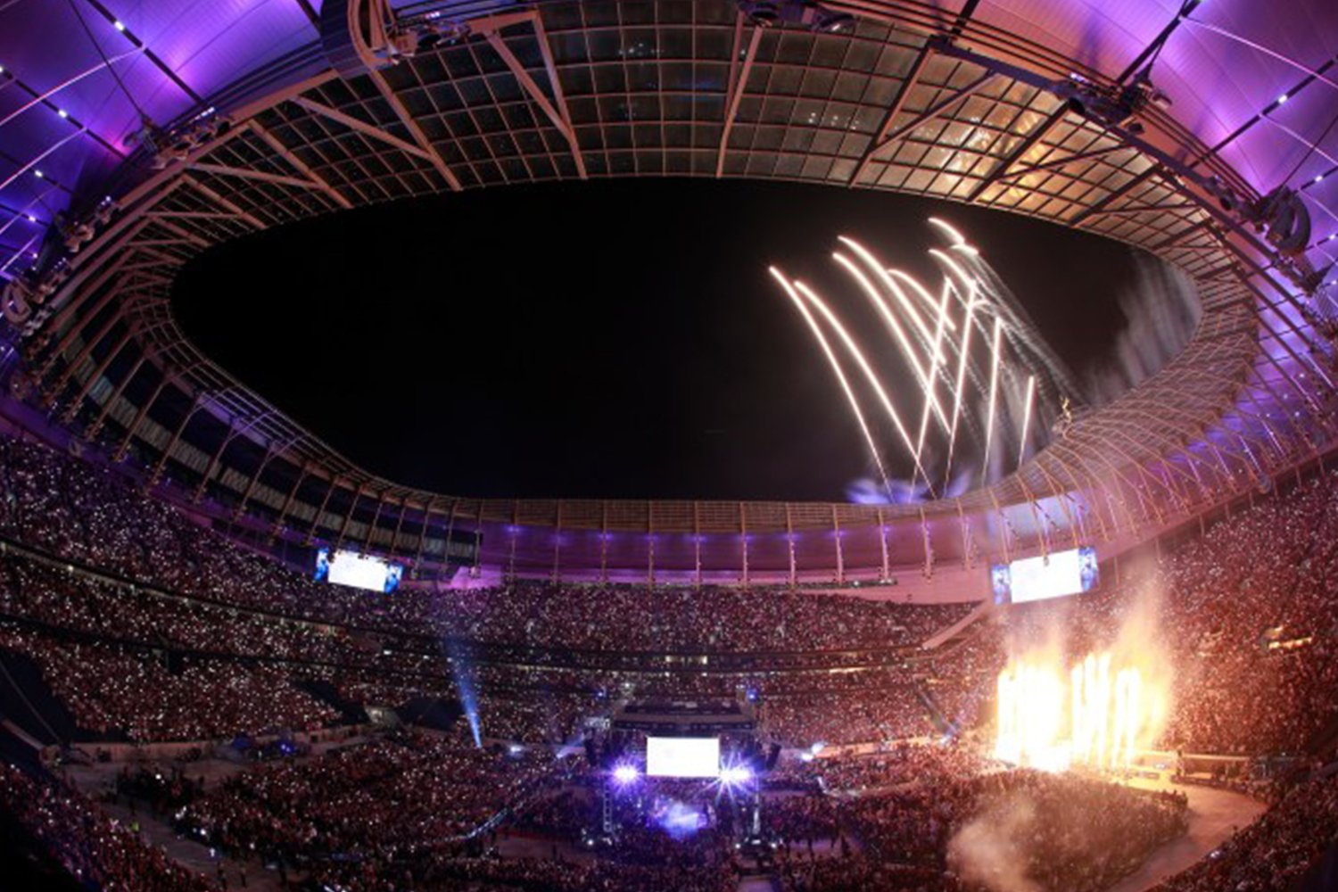 A large stadium filled with spectators at night, with fireworks and bright lights illuminating the sky and the stage.