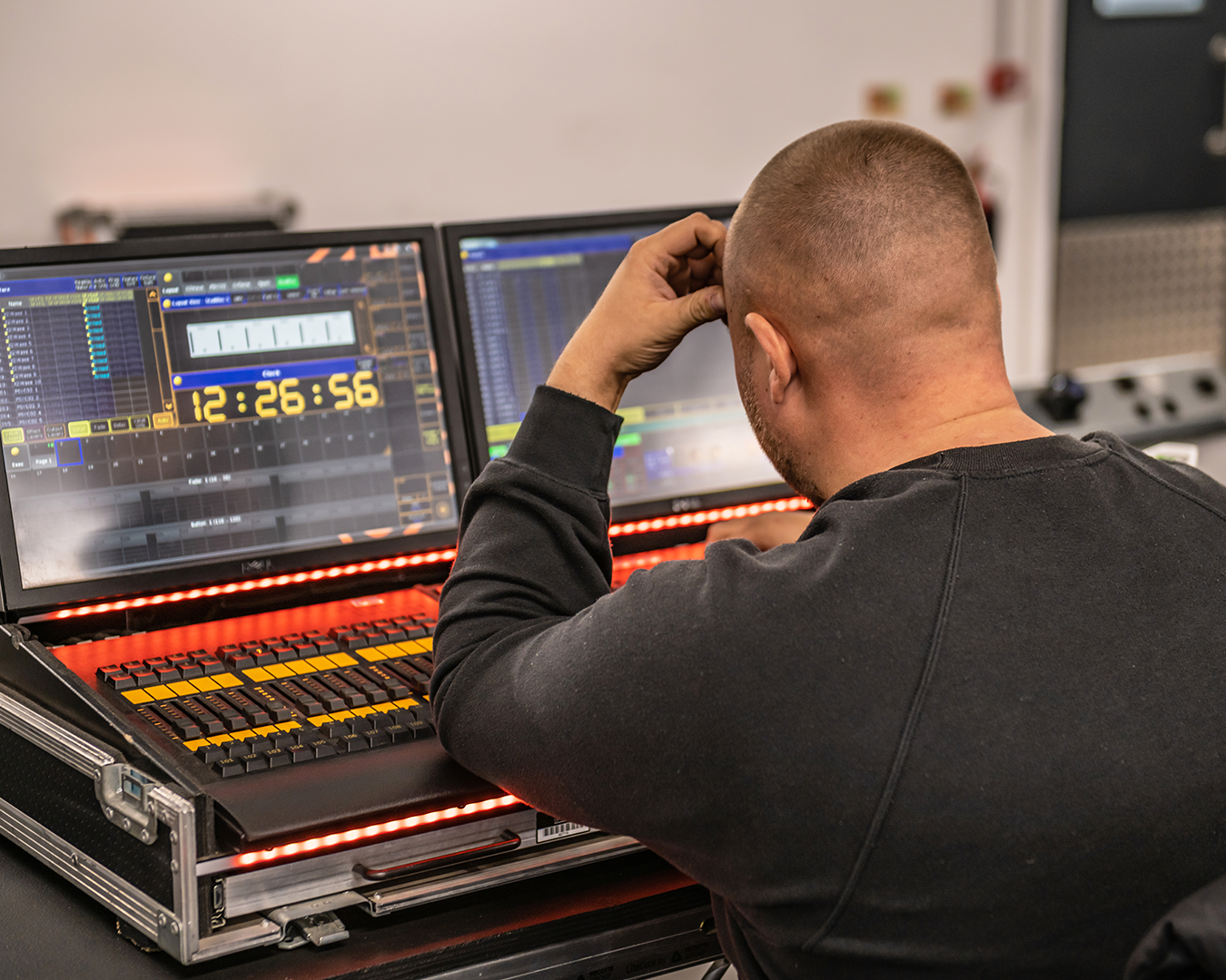 A man with a shaved head is working on music production using dual computer monitors displaying digital audio workstations, with a control surface and a digital timer showing 12:26:56.