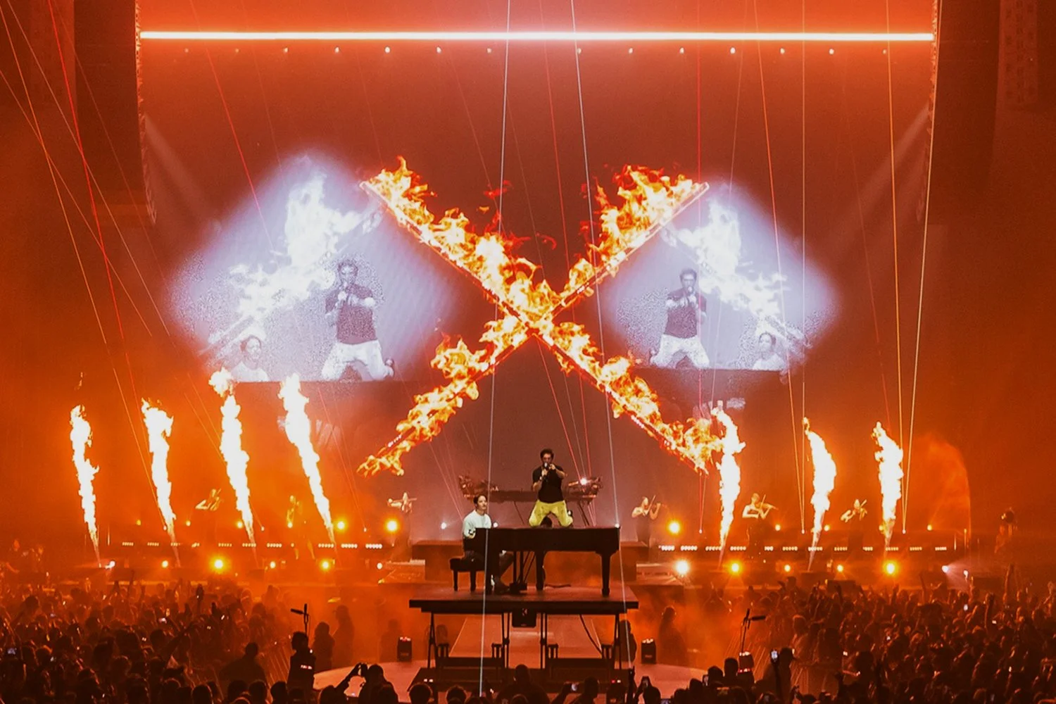 A concert stage with performers, pyrotechnics, and flames, with a large LED screen displaying images of performers, and an audience in the foreground.