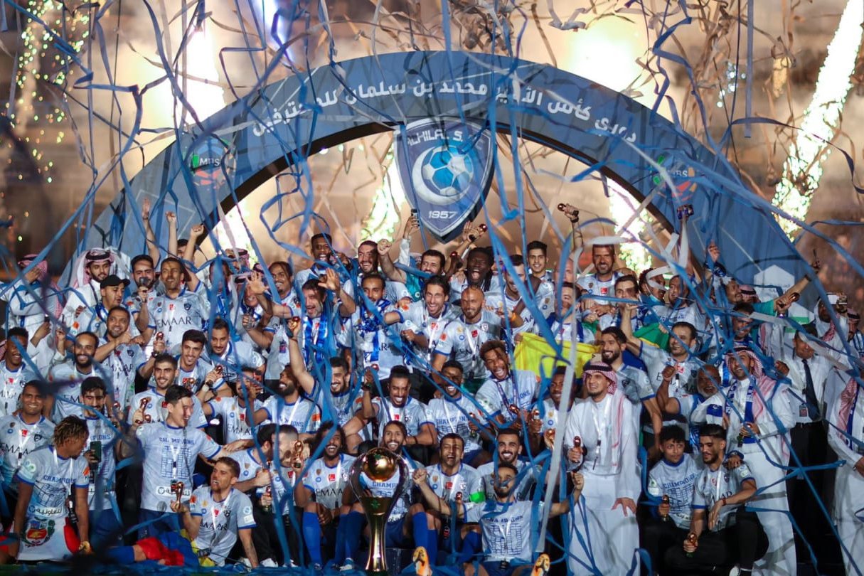 Group of soccer players celebrating with trophies, confetti, and streamers, under a large arch with a team emblem and Arabic script.