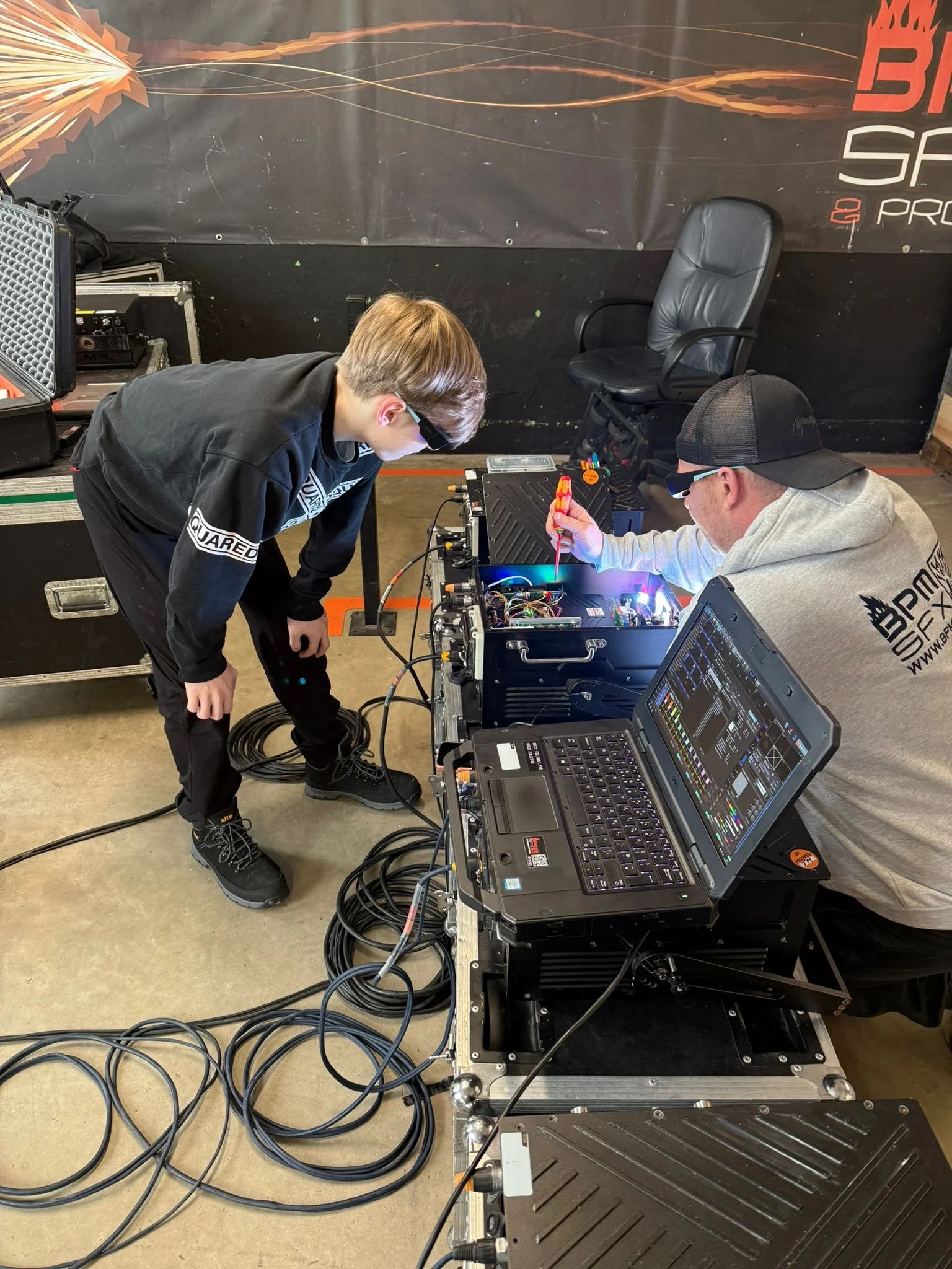 Two men working on electronic equipment at a table with a laptop, wires, and tools, in a workshop or event setting. One man is bending over, and the other is seated, wearing glasses and a cap.