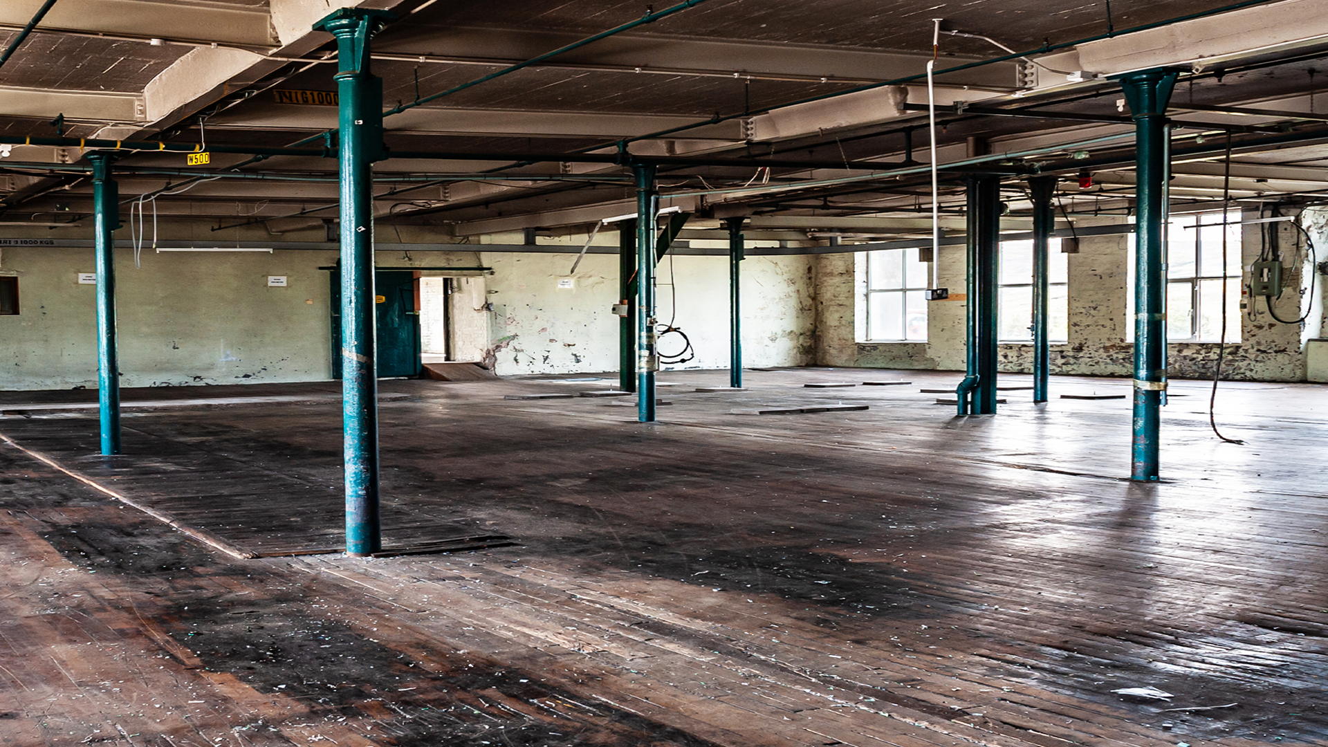 Empty, dilapidated warehouse with wooden flooring, green support columns, large windows, and peeling paint on the walls.
