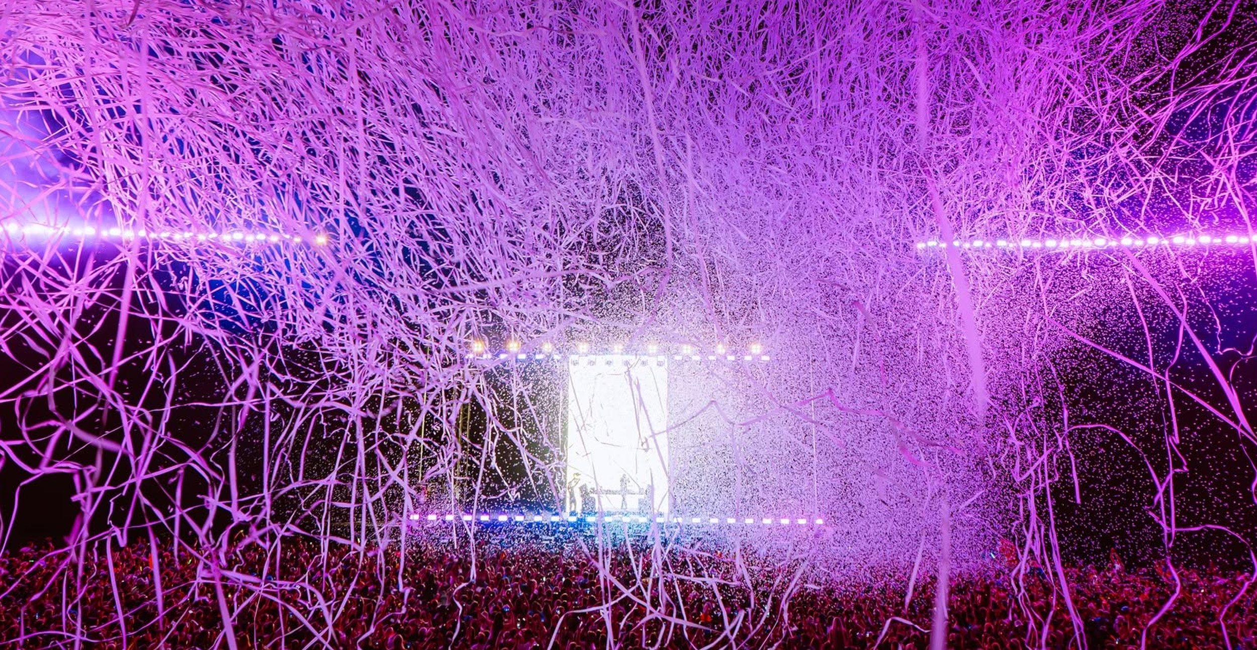 A large crowd gathered at night under a canopy of purple-lit tree branches with confetti falling and a stage in the distance.