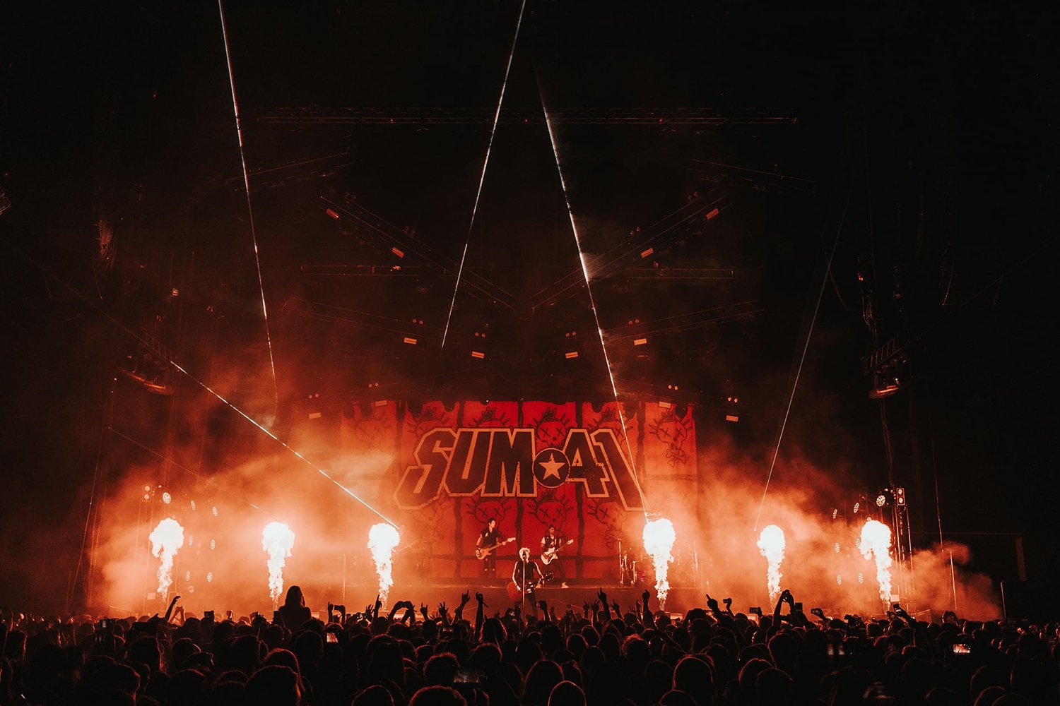 A live concert with a band performing on stage, illuminated by orange and red lighting, pyrotechnics, and smoke, with an audience in front taking photos and videos. The backdrop displays the word 'SUM41'.