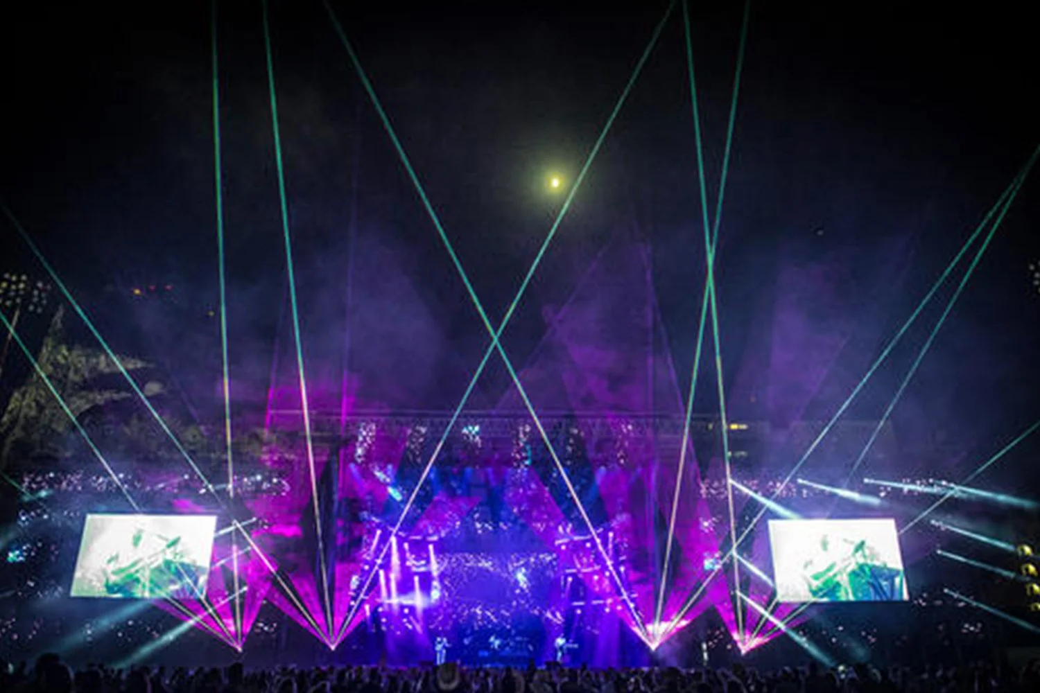 A concert scene at night with colorful laser lights, large screens displaying visuals, and a crowd of people.