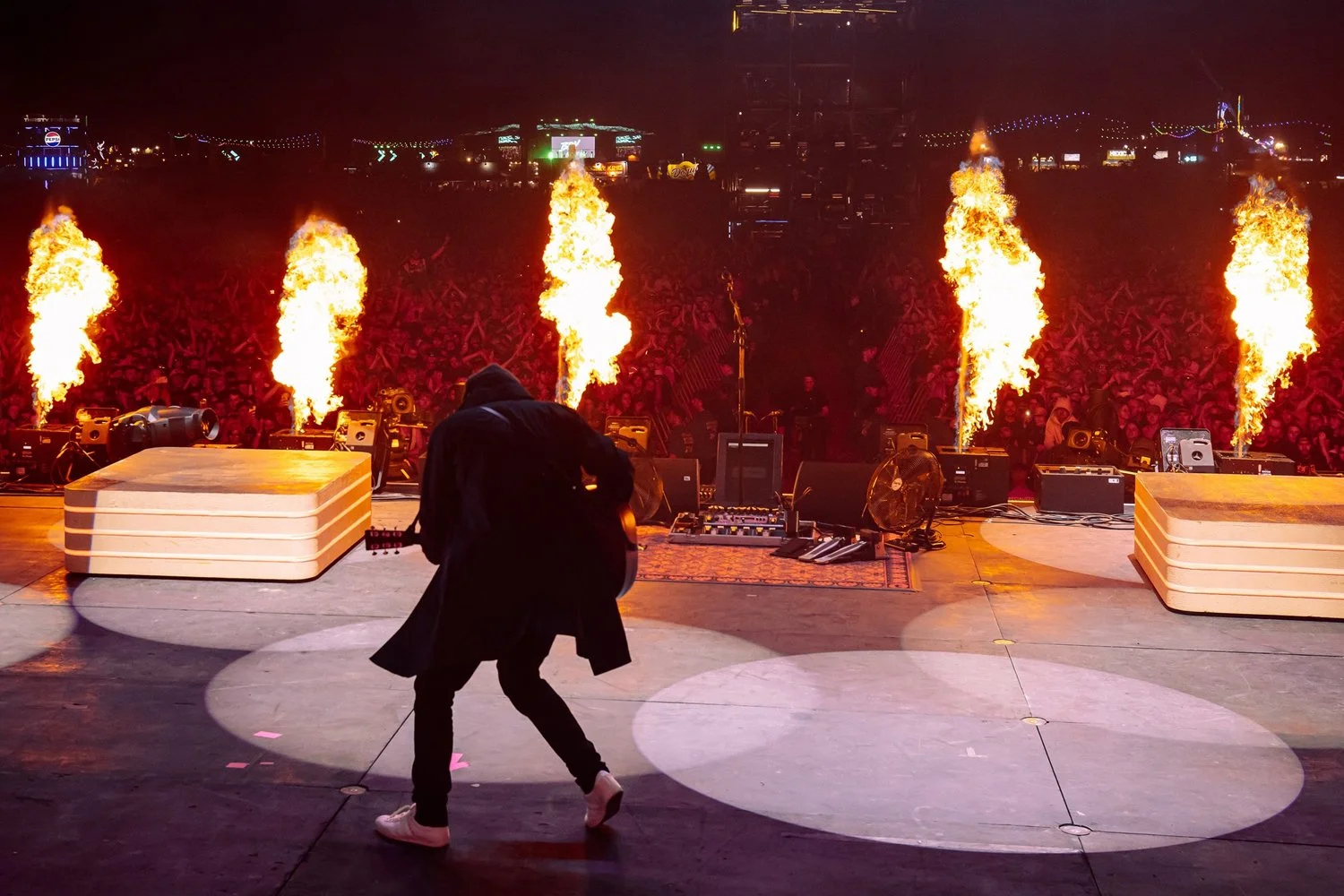 Musician performing on stage at night with flames shooting from pyrotechnics, crowd with raised hands in background, stage equipment and lighting visible.
