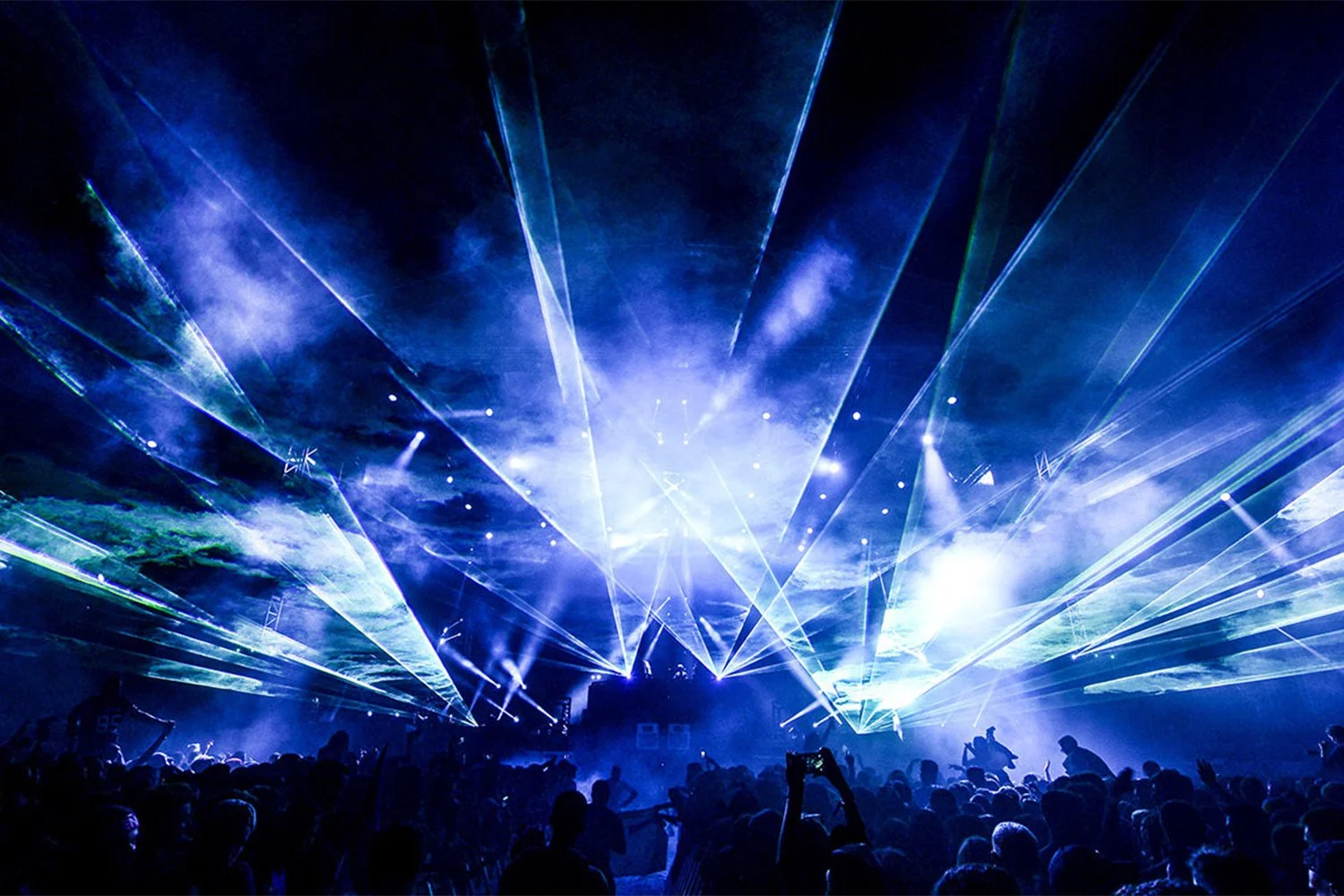 A concert scene with blue and white laser lights illuminating a crowd of people at night.