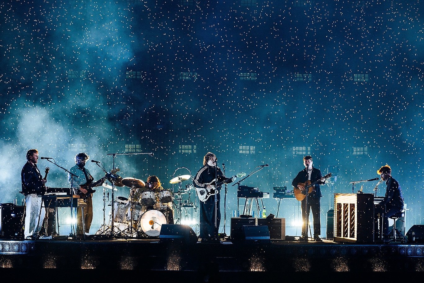 A band of six musicians performing on a stage with a starry night backdrop and blue lighting.