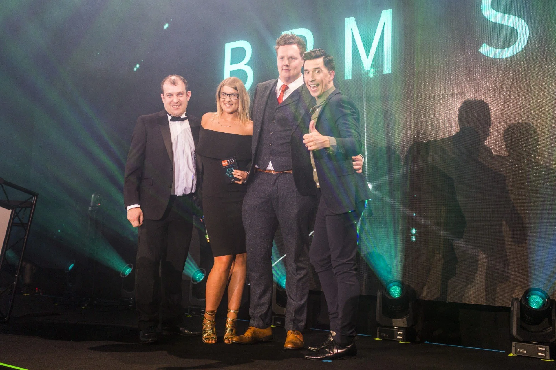 Four people dressed in formal attire standing on a stage with green and blue lighting, with large illuminated letters in the background spelling B R M.