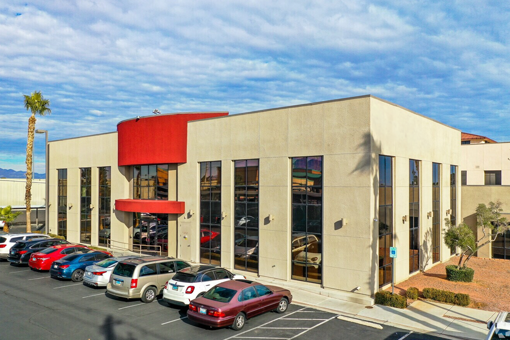 Modern commercial building with red accents, large glass windows, and a parking lot with several cars, including sedans and SUVs, under a partly cloudy sky.