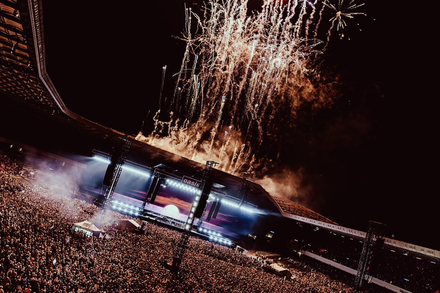 A large outdoor concert at night with fireworks lighting up the sky above the stage filled with a dense crowd of people.