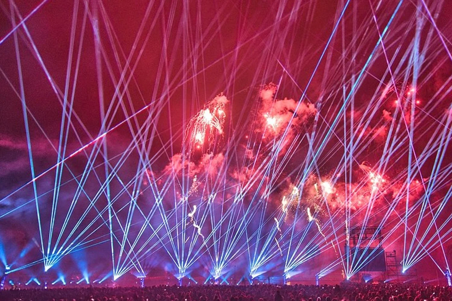 Colorful fireworks display with purple, red, and pink explosions and blue laser lights over a crowd at night.