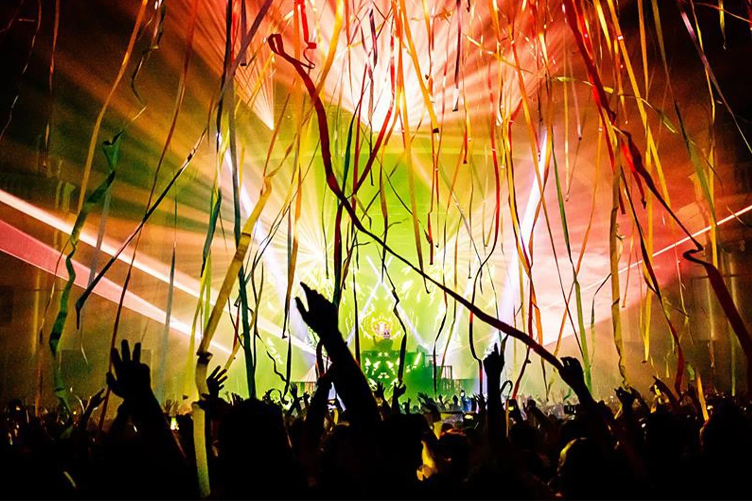 Crowd at a lively concert or dance event with streamers hanging from the ceiling, colorful lighting, and a DJ or performer on stage in the background.